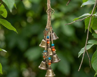 May include: A hanging wind chime featuring multiple brass-colored metal bells suspended by jute twine. The arrangement includes a decorative skeleton key, small metal charms shaped like a leaf, sun, and tree, and colorful beads in blue and orange.