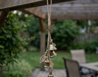 May include: A hanging wind chime featuring a bronze-toned butterfly ornament at the top, connected by natural jute twine to four small brass-colored bells and two metal leaf-shaped charms, suspended from a thick, braided rope loop for easy display.