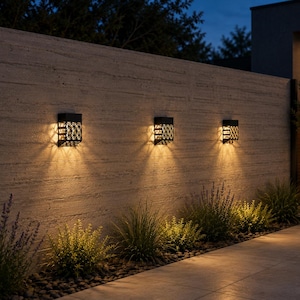 Puede incluir: Tres apliques de pared rectangulares de color negro para exteriores con un patrón de celosía decorativo. Cada lámpara emite un brillo cálido hacia abajo, iluminando la pared de piedra texturizada y el suelo con una luz suave y ambiental.
