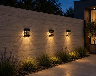 May include: Three rectangular black wall-mounted outdoor lights with a decorative lattice pattern. Each fixture emits a warm, downward-facing glow that illuminates the textured stone wall and the ground below in a soft, ambient light.