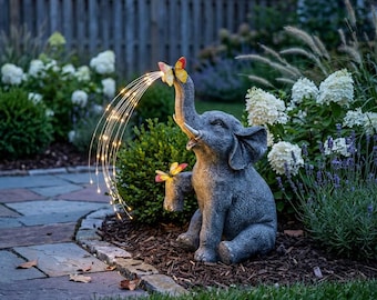May include: A gray elephant garden statue featuring a trunk raised upward with a spray of warm white LED fairy lights and a colorful butterfly perched on the tip, plus a second butterfly resting on the elephant's raised front foot.