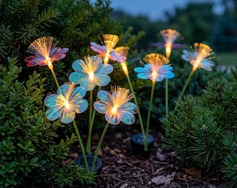 May include: A set of solar-powered garden lights shaped like flowers with iridescent petals and fiber optic centers that glow with warm yellow light. The lights are mounted on thin green stems and are positioned in dark mulch near green shrubs.