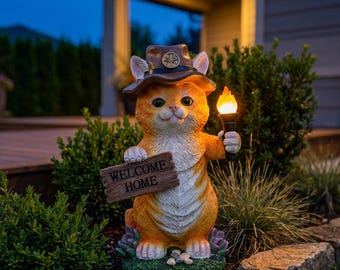 May include: An orange tabby cat garden statue wearing a brown hat with a compass emblem. The cat holds a wooden sign that reads WELCOME HOME and a torch with a glowing flame. The base features small green succulents and grey stones.