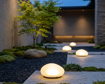 May include: A series of smooth, rounded, pebble-shaped outdoor lights emitting a warm, soft glow. These low-profile, dome-shaped lamps are positioned along a stone pathway, illuminating the surrounding dark gravel and patches of green moss.