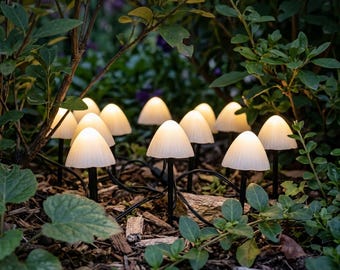 May include: A string of outdoor mushroom-shaped lights featuring white, ribbed, dome-shaped caps that emit a soft glow. The lights are connected by a thin black wire and are positioned among green foliage and wood chips in a garden setting.