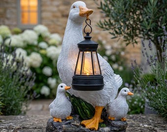 May include: A white goose figurine with orange feet and beak, holding a black lantern in its beak. Two smaller white ducklings with orange feet and beaks stand on a dark, textured base at the feet of the larger goose. The lantern emits a warm light.