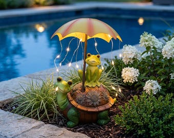 May include: A decorative garden figurine featuring a green turtle and a green frog sitting together under a yellow umbrella. The turtle shell is textured with sunflower patterns. Thin, illuminated wire strings hang from the umbrella edges.