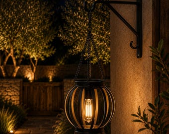 May include: A black metal spherical lantern with vertical slats, suspended by a matching chain from a wall-mounted bracket. The lantern contains a single glowing Edison-style light bulb, casting a warm light against the exterior wall.