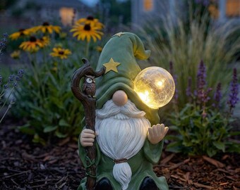 May include: A garden gnome figurine wearing a green hat with yellow stars, holding a brown staff topped with a small owl. The gnome has a long white beard and holds a glowing, spherical crackled glass light in the other hand.