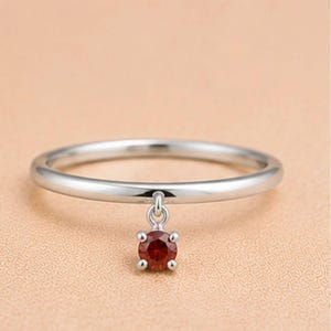 May include: A thin, polished silver-tone metal ring featuring a single, small, round-cut dark red gemstone charm suspended from the band by a tiny circular link. The gemstone is secured in a four-prong setting, creating a delicate, minimalist look.