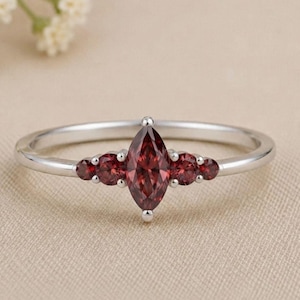 May include: A silver ring featuring a central marquise-cut deep red gemstone flanked by two smaller round-cut red stones on each side. The band is smooth and polished, showcasing a symmetrical design with prong settings for all five stones.