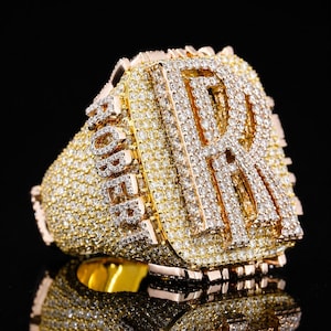 Op de afbeelding: Een grote gouden ring met een prominente letter R en de naam ROBERT gespeld in heldere, sprankelende stenen. Het gehele oppervlak van de ring is dicht bezet met kleine, ronde, briljant geslepen stenen, wat zorgt voor een sterk reflecterend, getextureerd uiterlijk op de gouden basis.