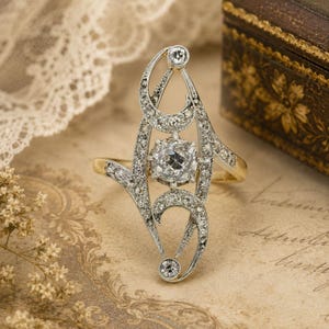 May include: A silver-toned metal ring featuring an elongated, openwork design set with numerous small, clear, sparkling stones. A larger, round, faceted clear stone is positioned in the center, with two smaller round stones at the top and bottom.