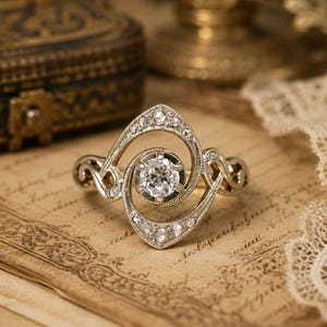 May include: A silver-toned ring featuring a central round-cut clear gemstone surrounded by a swirling, openwork metal frame. The band and the surrounding frame are embellished with multiple small, round-cut clear stones set into the metal.