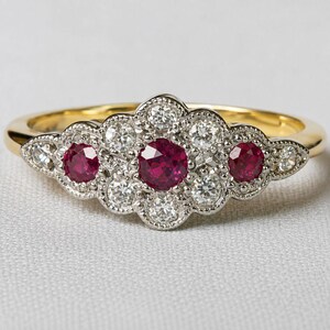 May include: A gold ring featuring a central cluster design with three round red rubies and eight round white diamonds. The stones are set in a silver-toned metal setting with milgrain detailing along the edges, creating a floral-like arrangement.