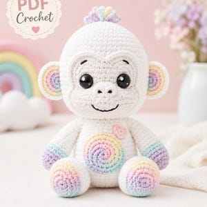 May include: A white crochet monkey doll featuring a rainbow-colored spiral on its belly, rainbow-colored hands, feet, and inner ears, a small pink heart patch on its chest, and a tuft of rainbow yarn on its head. The text PDF Crochet is visible.