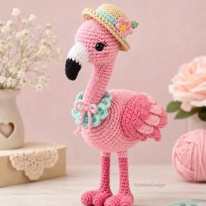May include: A crocheted flamingo toy in shades of pink, featuring a black and white beak, a light blue ruffled collar, and a beige straw-style hat adorned with a small pink flower. The text StichNestDesign is visible on the lower right side.