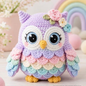 May include: A crochet owl plush toy featuring a lavender body with pastel pink, yellow, mint green, and blue scalloped feathers. It has large white and blue eyes, a yellow beak, and pink cheeks. A cluster of small crocheted flowers sits on its head.