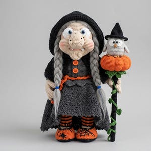 May include: A crocheted witch doll with gray braided hair, wearing a black hooded cloak and dress with orange buttons. The doll holds a staff topped with a small gray owl and an orange pumpkin. It wears orange and black striped socks and shoes.