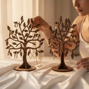 May include: Two dark brown wooden jewelry stands shaped like trees with multiple branches and leaves. Each stand features a circular base and numerous small holes designed for hanging earrings, necklaces, and other jewelry items.