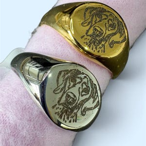 May include: Two signet rings, one in a polished gold tone and one in a polished silver tone, each featuring a detailed laser-engraved portrait of a dog on the circular face. The rings are displayed on a soft, light pink cylindrical holder.