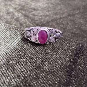 Può includere: Un anello color argento con una pietra preziosa magenta di forma ovale al centro. La fascia in metallo è decorata con dettagliate incisioni a forma di foglia su entrambi i lati della pietra, creando un aspetto strutturato su tutta la superficie.