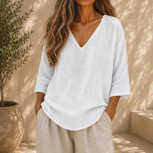 May include: A white long-sleeve blouse featuring a deep V-neckline and a loose, relaxed fit. The garment is crafted from a textured fabric and is paired with light beige trousers. The design includes three-quarter length sleeves and a straight hem.