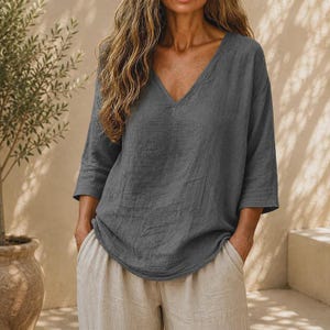 May include: A charcoal gray long-sleeve top with a V-neckline and a loose, relaxed fit. The garment features three-quarter length sleeves and a straight hemline, paired with light beige trousers that have side pockets and a comfortable, wide-leg cut.