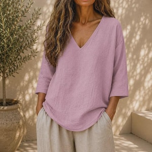 May include: A light mauve V-neck top with three-quarter length sleeves and a loose, relaxed fit. The garment features a textured fabric finish and is paired with light beige trousers, creating a simple, solid-colored ensemble for daily wear.