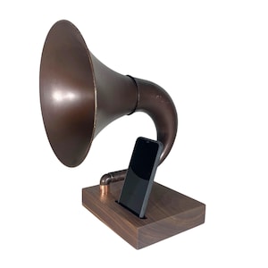 May include: A vintage-style phone speaker with a copper-colored horn and a dark wooden base. A black smartphone is placed in a slot on the base. The speaker has a classic gramophone design, blending retro aesthetics with modern technology.