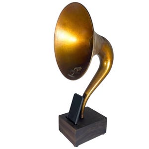 May include: A vintage-style brass gramophone speaker with a dark wooden base. The speaker has a large, flared horn and a curved neck. A smartphone is placed in a slot in the base. The speaker is designed to amplify sound.