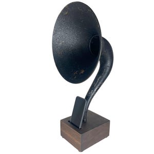 May include: A black, vintage-style gramophone-shaped phone holder. The horn is large and circular, with a curved arm connecting it to a wooden base. A black phone is inserted into a slot in the base.