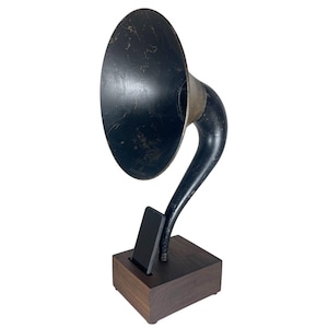 May include: A black and gold-toned gramophone-shaped phone holder. The horn is large and oval, with a curved neck leading to a wooden base. A black phone is inserted into the base, creating a vintage aesthetic.