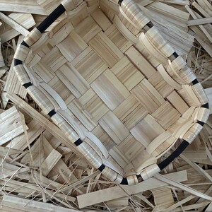 Puede incluir: Una cesta rectangular tejida con tiras de madera natural de color beige claro entrelazadas con un marco negro oscuro. La cesta tiene una base plana y lados verticales construidos con un patrón de tejido simple, mostrando la textura de la madera.