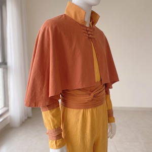 K&ouml;nnte beinhalten: Ein rostorangefarbenes Cape mit hohem Kragen und drei Knebelverschl&uuml;ssen &uuml;ber einem gelben Langarmshirt und einer passenden gelben Hose. Eine breite, rostorangefarbene Sch&auml;rpe ist um die Taille gewickelt und passende B&auml;nder akzentuieren die Handgelenke.