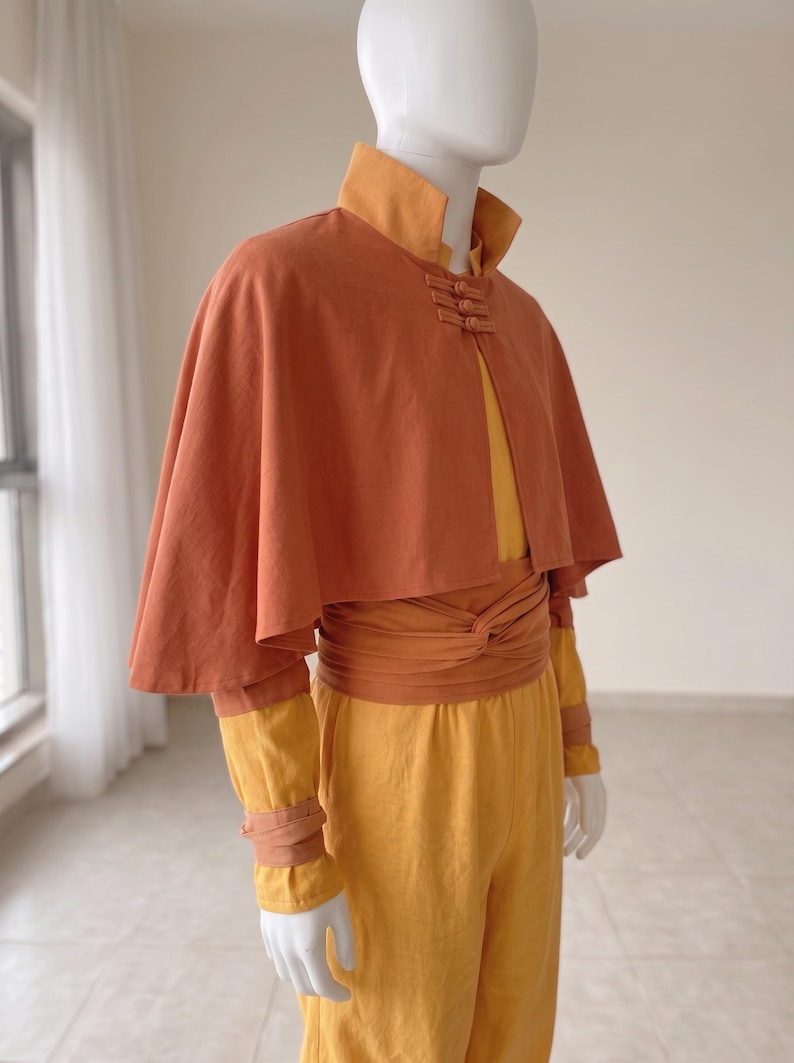 K&ouml;nnte beinhalten: Ein rostorangefarbenes Cape mit hohem Kragen und drei Knebelverschl&uuml;ssen &uuml;ber einem gelben Langarmshirt und einer passenden gelben Hose. Eine breite, rostorangefarbene Sch&auml;rpe ist um die Taille gewickelt und passende B&auml;nder akzentuieren die Handgelenke.