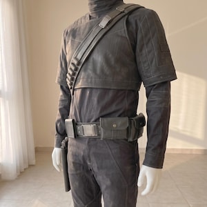 Pu&ograve; includere: Un set di costume tattico grigio scuro composto da una maglia a maniche lunghe con collo alto, uno strato superiore a maniche corte con imbottitura sulle spalle, una cintura multiuso con tasche e fibbia metallica e una tracolla diagonale.