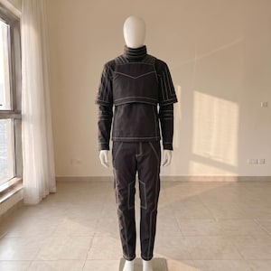Pu&ograve; includere: Un completo a due pezzi grigio antracite scuro composto da una maglia a maniche lunghe con collo alto e un pannello pettorale sovrapposto, abbinato a pantaloni coordinati. L'intero capo presenta evidenti cuciture bianche a vista.