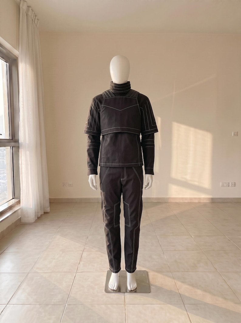 Pu&ograve; includere: Un completo a due pezzi grigio antracite scuro composto da una maglia a maniche lunghe con collo alto e un pannello pettorale sovrapposto, abbinato a pantaloni coordinati. L'intero capo presenta evidenti cuciture bianche a vista.