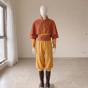 K&ouml;nnte beinhalten: Ein Kost&uuml;mset bestehend aus einer gelben lang&auml;rmeligen Tunika und passender gelber Hose, kombiniert mit einem rostorangen Cape mit Stehkragen und Knebelverschluss. Eine breite rostorange Sch&auml;rpe ist um die Taille gebunden, dazu dunkelbraune Beinwickel.