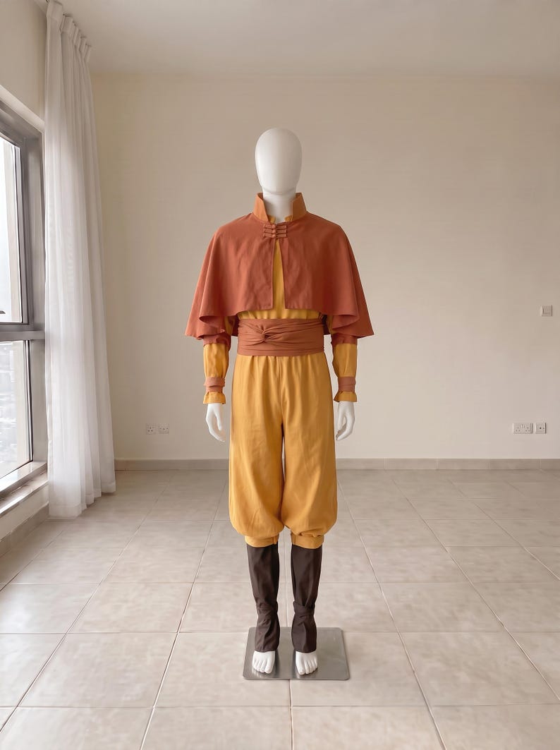 K&ouml;nnte beinhalten: Ein Kost&uuml;mset bestehend aus einer gelben lang&auml;rmeligen Tunika und passender gelber Hose, kombiniert mit einem rostorangen Cape mit Stehkragen und Knebelverschluss. Eine breite rostorange Sch&auml;rpe ist um die Taille gebunden, dazu dunkelbraune Beinwickel.