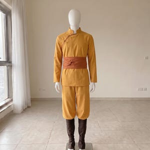 K&ouml;nnte beinhalten: Eine senfgelbe Tunika mit langen &Auml;rmeln, Stehkragen und seitlichen Knebelkn&ouml;pfen, kombiniert mit einer passenden senfgelben Hose und einer breiten, rostorangen Sch&auml;rpe in der Taille sowie dunkelbraunen Beinwickeln an den Unterschenkeln.
