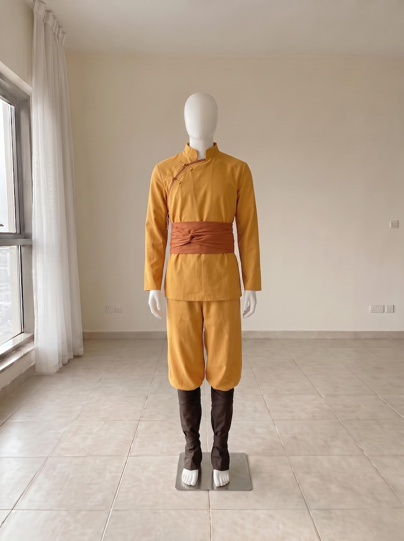 K&ouml;nnte beinhalten: Eine senfgelbe Tunika mit langen &Auml;rmeln, Stehkragen und seitlichen Knebelkn&ouml;pfen, kombiniert mit einer passenden senfgelben Hose und einer breiten, rostorangen Sch&auml;rpe in der Taille sowie dunkelbraunen Beinwickeln an den Unterschenkeln.