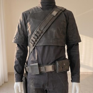 Pu&ograve; includere: Un set di costume tattico grigio scuro composto da una maglia a maniche lunghe con collo alto, un gilet a maniche corte e pantaloni abbinati. L'outfit include una tracolla diagonale con passanti, una cintura con tasche e una fondina.