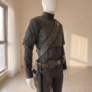 Pu&ograve; includere: Un costume grigio scuro su un manichino, composto da un top a maniche corte con collo alto, una maglia a maniche lunghe e pantaloni abbinati. Una cintura nera con fondina e una tracolla diagonale con alloggiamenti per munizioni.