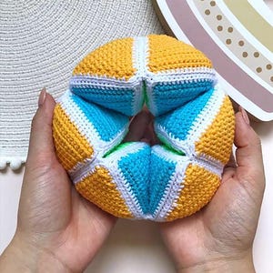 Op de afbeelding: Een bolvormig puzzelspeeltje opgebouwd uit gehaakte segmenten in afwisselend felgeel en levendig blauw. De segmenten zijn verbonden door witte gehaakte naden, wat een gestructureerd, geometrisch patroon vormt in een ronde vorm.