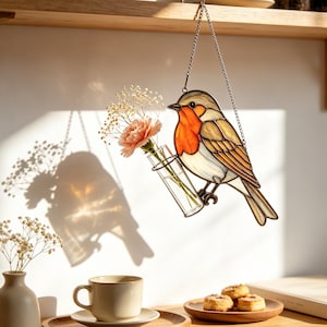 Puede incluir: Un atrapasoles de vidrieras con forma de petirrojo, con pecho naranja y alas marrones, suspendido por una cadena metálica. El pájaro sostiene un pequeño jarrón de tubo de ensayo de cristal con un clavel rosa y ramas de paniculata.