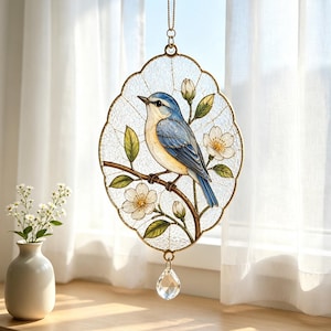 May include: An oval-shaped stained glass suncatcher featuring a blue and cream-colored bird perched on a brown branch with white flowers and green leaves. The piece has a gold-toned metal frame and a clear, faceted crystal drop at the bottom.