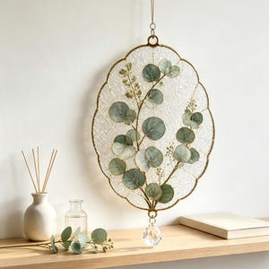 May include: An oval wall hanging featuring a gold metal frame with a delicate white lace-like mesh interior. Pressed green eucalyptus leaves are arranged inside the frame, and a clear faceted crystal drop hangs from the bottom centre.