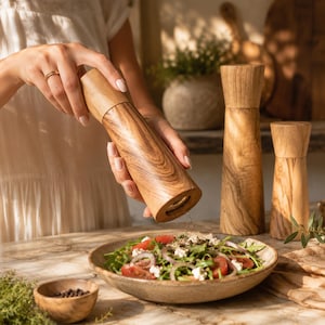 Op de afbeelding: Een hoge, cilindrische houten pepermolen met een zichtbaar natuurlijk nerfpatroon. De molen wordt schuin gehouden boven een keramische kom gevuld met een salade van gesneden tomaten, rode uien en witte kaas, met twee extra molens in de buurt.